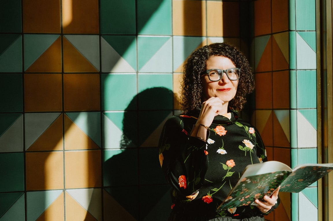 A woman with curly hair and glasses stands in front of a colorful tiled wall, smiling and holding an open book. She’s wearing a dark floral top and is bathed in warm sunlight streaming through a window.