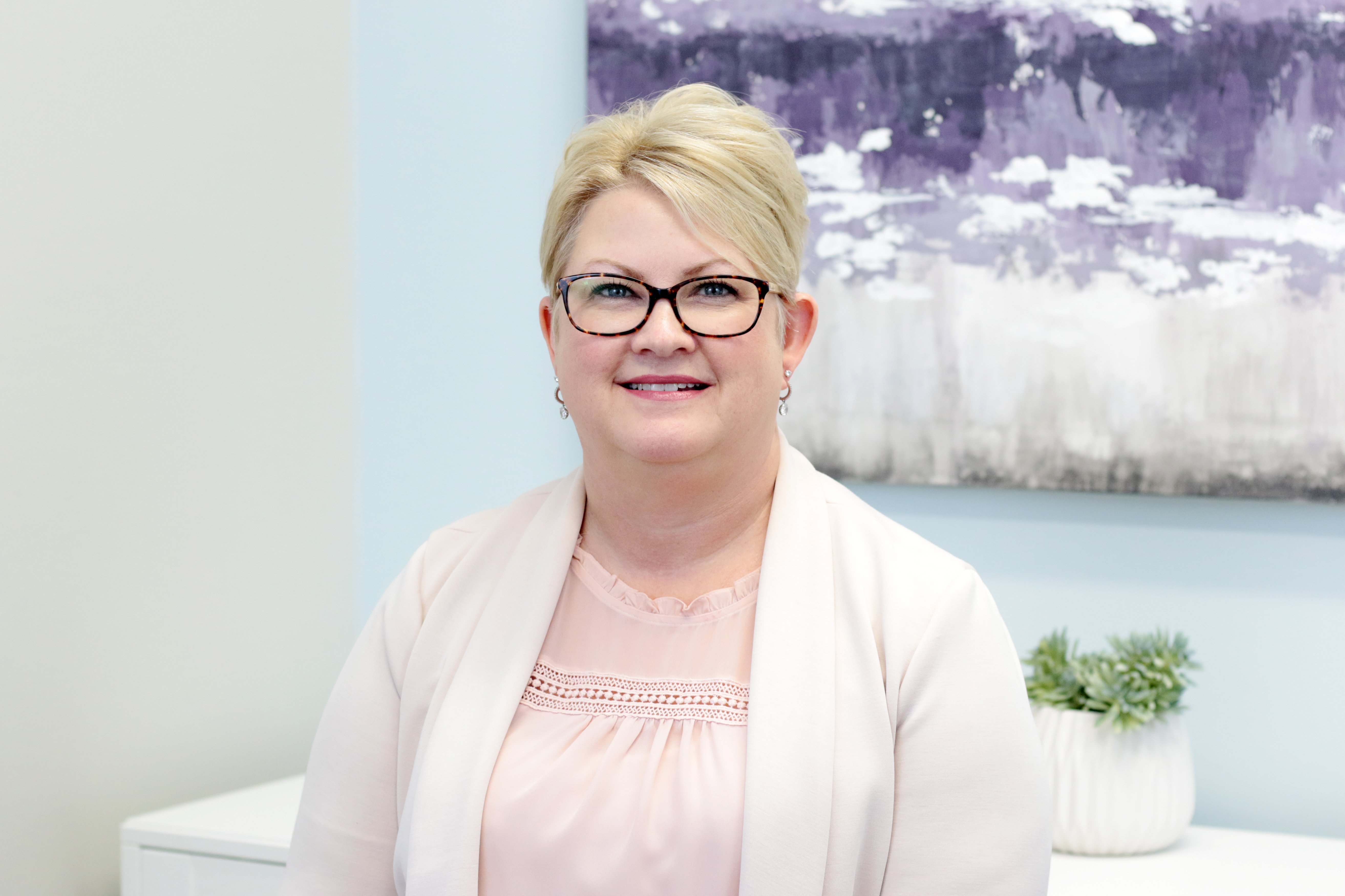 Larissa Warne, a smiling woman with short blond hair and glasses, wearing a light pink blouse and cream jacket, seated in front of a purple abstract painting and a small plant.